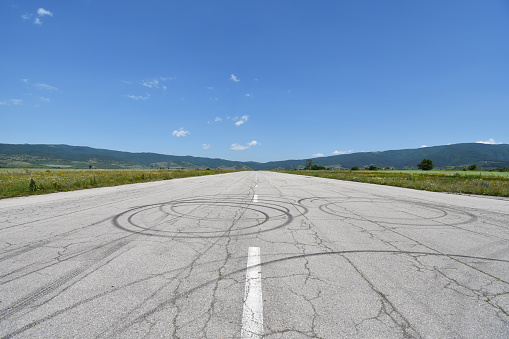 Reaching the horizon deserted military airport runway near Sapareva Banya, Bulgaria, nowadays used for amateur car races, cracks and tire tracks seen on the old asphalt surface. Summer day with serene blue sky.