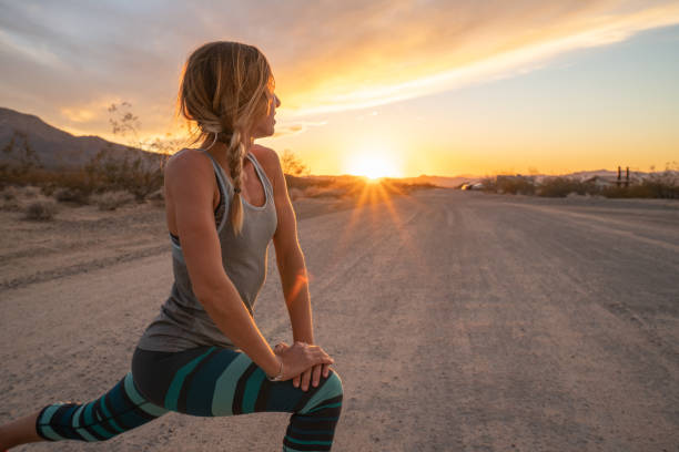 mulher nova que estica o corpo após movimentar-se, por do sol na extremidade da estrada; feminino estica o corpo na natureza - exercising running women jogging - fotografias e filmes do acervo