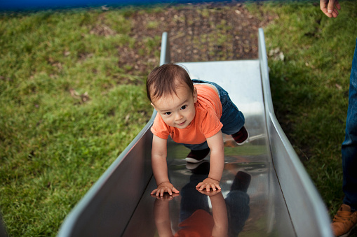 An adorable baby boy is climbing up a slide in the park looking happy and full of vitality.