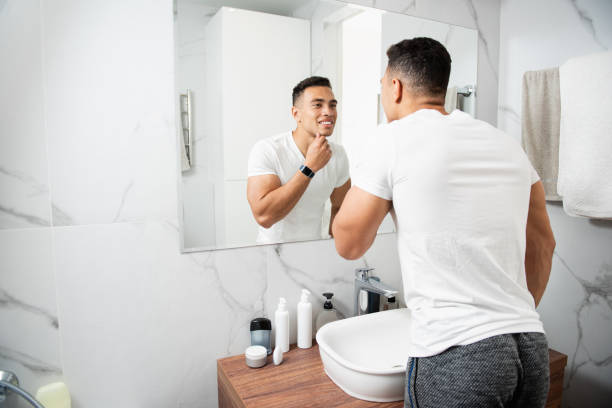 Smiling young man touching his chin before mirror Morning hygiene concept. Back side waist up portrait of young brunette muscular smiling guy in white t-shirt looking at reflection on mirror and touching his chin shaving stock pictures, royalty-free photos & images