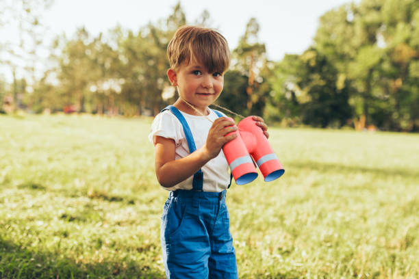 petit garçon jouant avec des jumelles recherchant une imagination ou l'exploration dans la journée d'été en stationnement. enfant heureux jouant le jeu de safari semblant dehors dans la forêt. concept d'enfance - discovery binoculars boy scout searching photos et images de collection