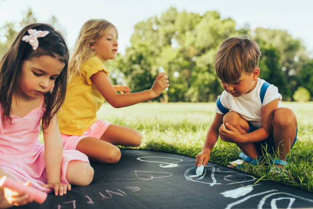 Outdoors image of kids sitting on the green grass playing with colorful chalks. Happy children drawing with chalks in the park. Friends, boy and two girls having fun on sunlight outdoor. Childhood stock photo