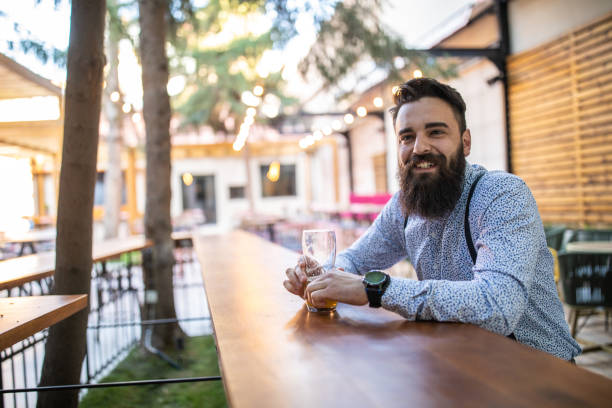 Cheerful Hipster man sitting in an open pub alone, having a few beers after long day at work, looking around and smiling happily