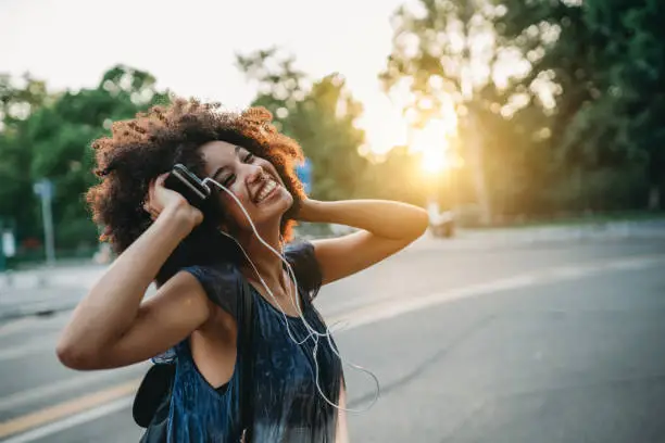 Photo of Young adult woman with afro hair dancing in the city at sunset while listening to music