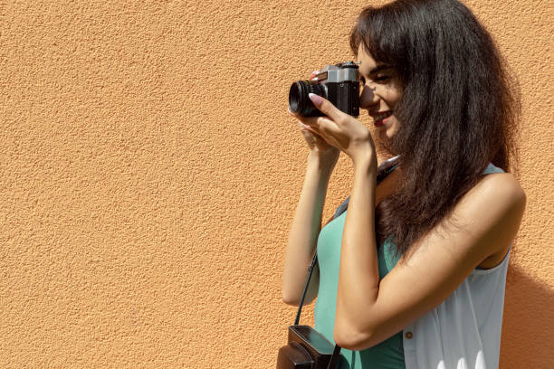 istock Beautiful young lady with retro camera standing against light orange wall 1157995603