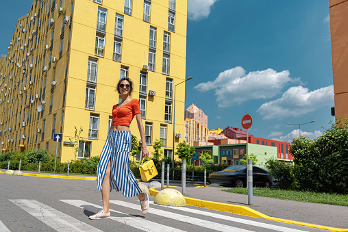 Full length portrait of charming brunette girl in long striped skirt walking on the street and using pedestrian crossing. She holding yellow purse and smiling