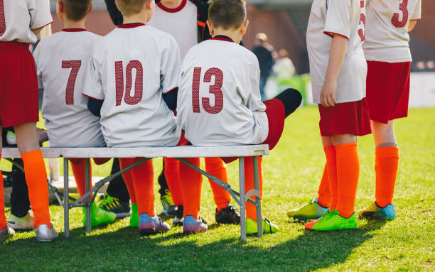 grupa dzieci siedzących na ławce sportowej na boisku szkoły trawiastej. chłopcy w piłce nożnej siedzą na ławce rezerwowych - soccer bench soccer player sport zdjęcia i obrazy z banku zdjęć