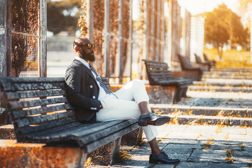 An elegant bald bearded black guy in sunglasses and the fashionable costume is sitting on a wooden bench in a public park and thoughtfully looking straight; a fancy African man outdoors, sunny day