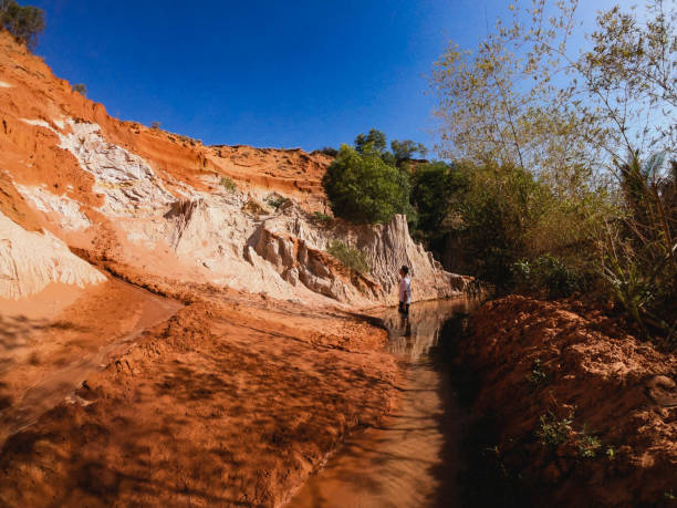A wide-view shot of a young male tourist standing in a stream of water while exploring the red rock canyon of Vietnam.