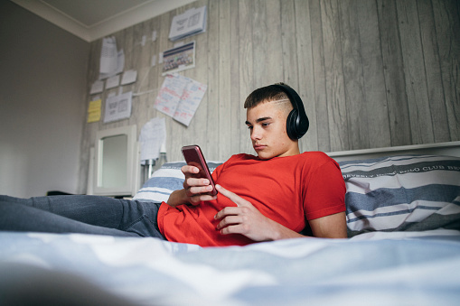 One Teenage boy only lying down on his bed relaxing. He is listening to music and using his smart phone while on social media.