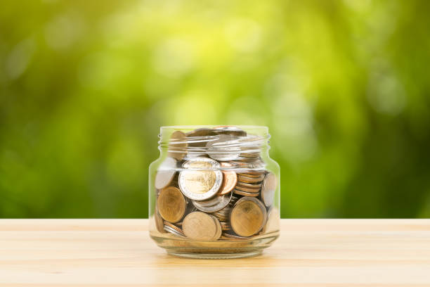 istock Coin glass jar container on wooden desk, saving concept, on green tree background 1158019551
