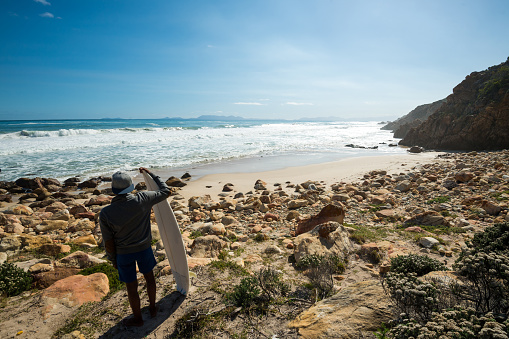 Rear view of teenage boy with surfboard standing at beach. Sporty male is looking at sea waves against sky. He is enjoying summer vacation.