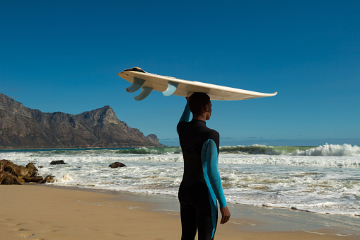 Rear view of confident teenage boy carrying surfboard on head. Surfer is wearing wetsuit during summer vacation. Male is looking at sea waves against blue sky.