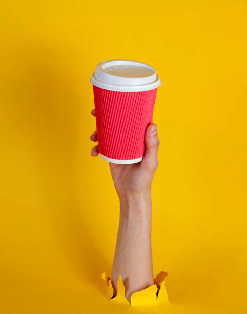 Female hand holds paper coffee cup through torn yellow paper. Minimalistic concept