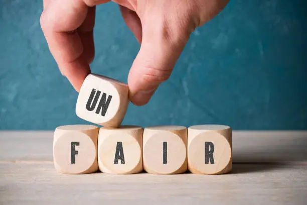 Unidentifiable person stacking blocks with black letters on side to spell unfair in front of scuffed blue wall