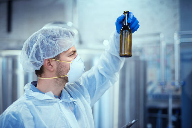istock Technologist in white protective suit with hairnet and mask working in food and beverage factory. Man specialist checking bottles for beverage production. 1158034353