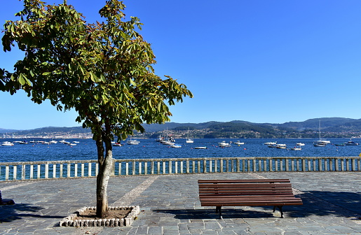 Rias Baixas, Pontevedra Province, Galicia, Spain. Bay with sailing boats and bench on a promenade. Blue sky, sunny day.