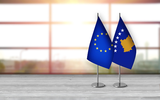 3D kosovan and european union flags with metallic pole, standing together on a white wooden desk in front of sunny window background. With large copy space you can write your own titles effectively. Also you can use this compositon as square in social media channels like instagram etc.