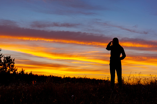 A woman looking at an amazing colorful sunset. Sky and cloud from dark blue through orange to yellow. You can see the contours of meadows and trees.