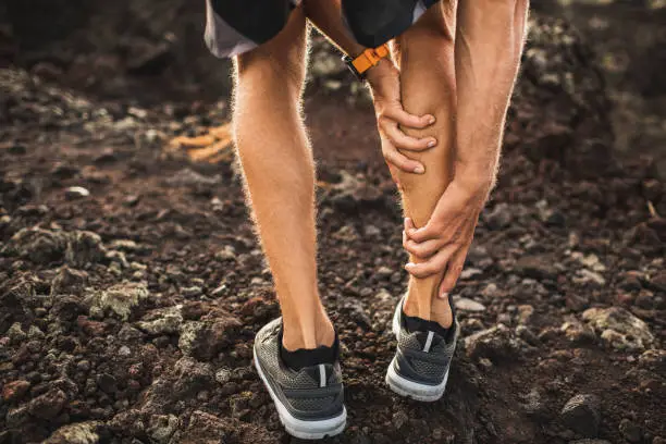 Male runner holding injured calf muscle and suffering with pain. Sprain ligament while running outdoors. View from the back close-up.