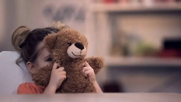 Sad female kid covering face by teddy bear toy, family problem, loneliness abuse