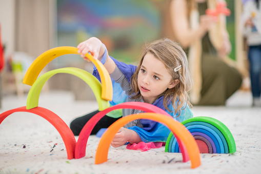 A girl places colourful rings on top of each other while staying incredibly focused. She is lying on the carpet of a classroom. She is dressed casually.