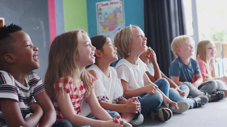 Group Of Laughing Elementary School Pupils Sitting On Floor Listening To Teacher