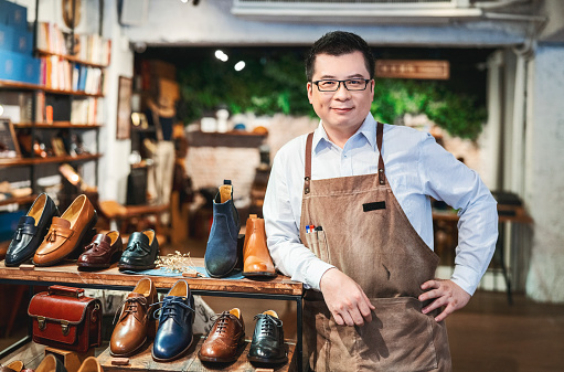 Portrait of confident male owner standing by shoe displayed on rack. Smiling entrepreneur with hand on hip in shoe store. He is wearing apron at workplace.