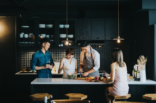 Group of joyful young Asian man and woman cooking together in the kitchen, chatting joyfully and enjoying red wine while preparing food for party