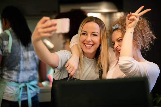 Taking a selfie making victory sign with her flatmate in the kitchen Cheerful young women taking a selfie with smart phone in the kitchen with flatmates in the back flatmate stock pictures, royalty-free photos & images