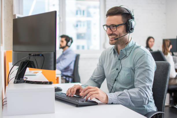 istock Smiling handsome male customer support phone operator with headset working in call centre. Group of sales agent working in office. 1158151781