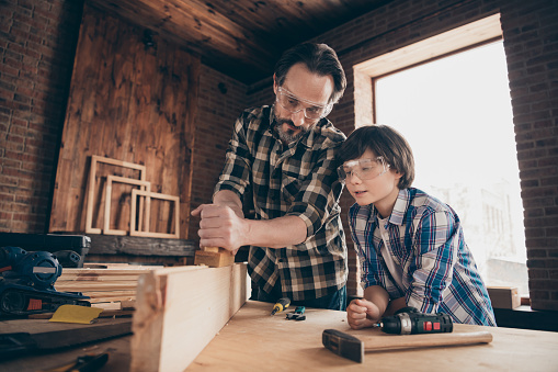 Portrait of focused serious pensive daddy son kid touch wood hold, hand equipment craftsman fix engineer plank bearded checkered shirt brunet hair interior indoors glasses goggles protective