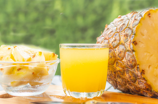 Sliced pineapple kept on a wooden table besides a glass filled with pineapple juice and a glass bowl with pineapple slices