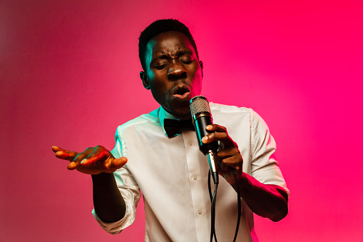 Young african-american jazz musician singing a song on gradient pink-red background in neon light. Concept of music, hobby, festival, open-air. Joyful guy improvising. Colorful retro portrait of singer.