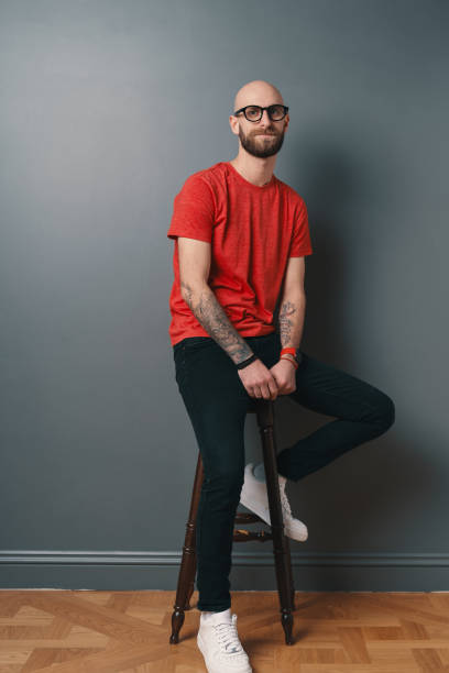 istock Young smiling man with beard and glasses on gray studio background while sitting on a tall chair. 1158169369