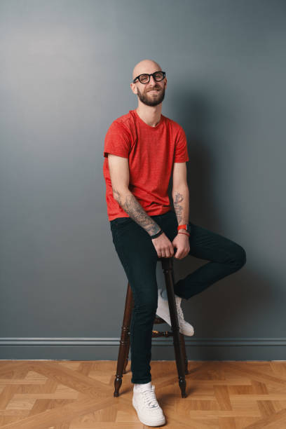 istock Young smiling man with beard and glasses sitting on a tall chair on gray studio background 1158169370