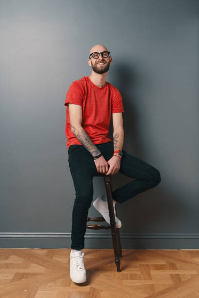 istock Young smiling man with beard and glasses sitting on a tall chair on gray studio background 1158169380