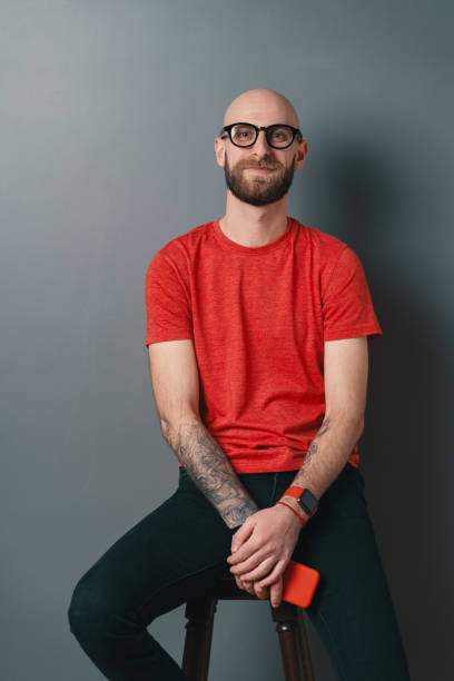 istock Young smiling man with beard and glasses sitting on a tall chair on gray studio background 1158169381