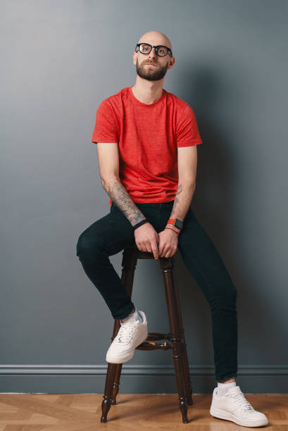 Confident young man posing in the studio on gray background while sitting on a tall chair stock photo