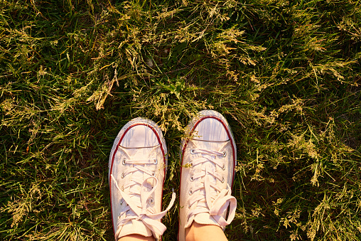 From above crop view of person in white sneakers standing on bright green grass in sunlight