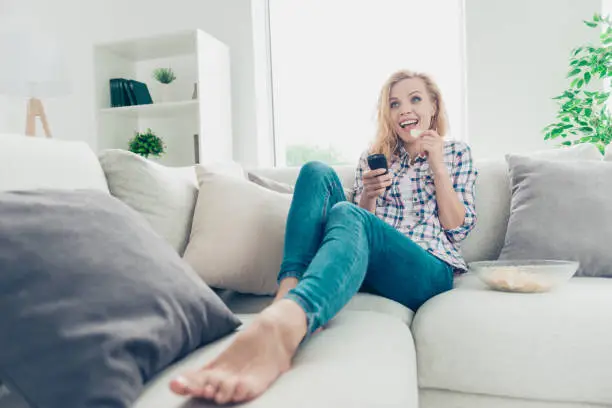 Low angle view of her she nice-looking attractive lovely charming, cute cheerful cheery glad wavy-haired girl in checked shirt lying on divan looking comedian at white light style interior living-room
