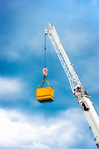 Crane boom with hook lifting a yellow container in harbor against blue sky.