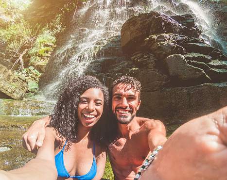 Happy couple taking selfie photo with smartphone camera under tropical waterfalls in summer vacation - Young people making photo souvenir - Focus on faces - Travel, youth and relationship concept