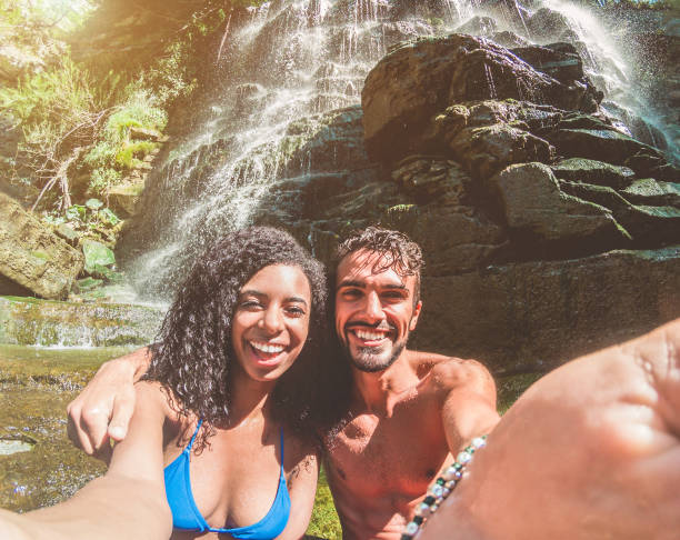 pares felizes que tomam a foto do selfie com a câmera do smartphone cachoeiras tropicais em férias de verão-jovens que fazem a lembrança da foto-foco nas faces-conceito do curso, da juventude e do relacionamento - river swimming women water - fotografias e filmes do acervo