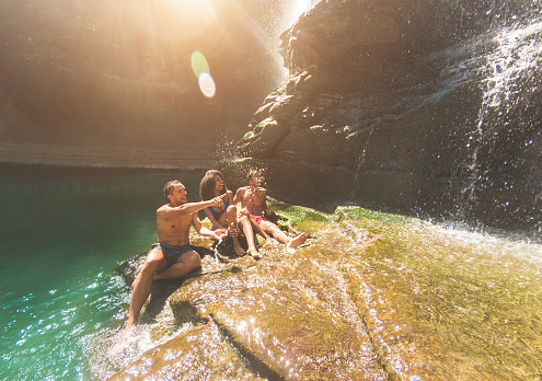 Group of friends having fun under waterfalls river - Young people swimming inside emerald water lagoon - Travel, summer and friendship concept - Focus on left guy face