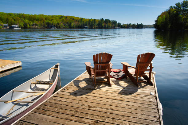 Muskoka chairs on a wooden dock Two Muskoka chairs on a wooden dock overlooking the blue water of a lake in Muskoka, Ontario Canada. A red canoe is tied to the pier and life jackets are visible near the chairs. boat dock lake stock pictures, royalty-free photos & images