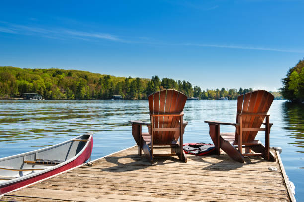 Muskoka chairs on a wooden dock Two Muskoka chairs on a wooden dock overlooking the blue water of a lake in Muskoka, Ontario Canada. A red canoe is tied to the pier and life jackets are visible near the chairs. summer stock pictures, royalty-free photos & images