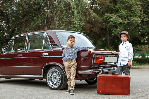 two Caucasian little boys in vintage clothes standing next to a retro car