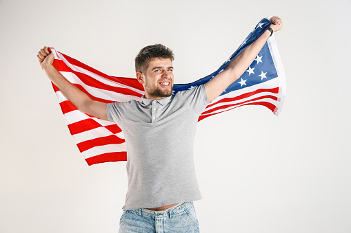 Celebrating an Independence day. Stars and Stripes. Young man with the flag of the United States of America isolated on white studio background. Looks crazy happy and proud as a patriot of his country.