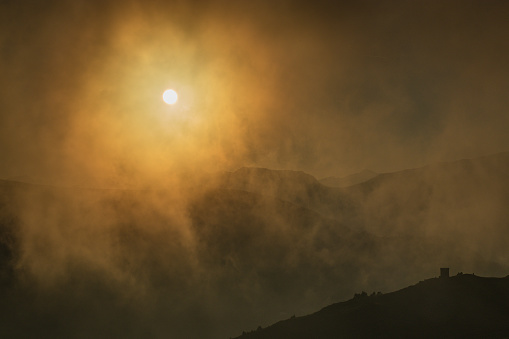 Sun hiding behind misty clouds on top of the mountain. golden sun shining through the mountains covered with fog, clouds or mist in evening hours. Silhouette of hills and mountains lit by sun.